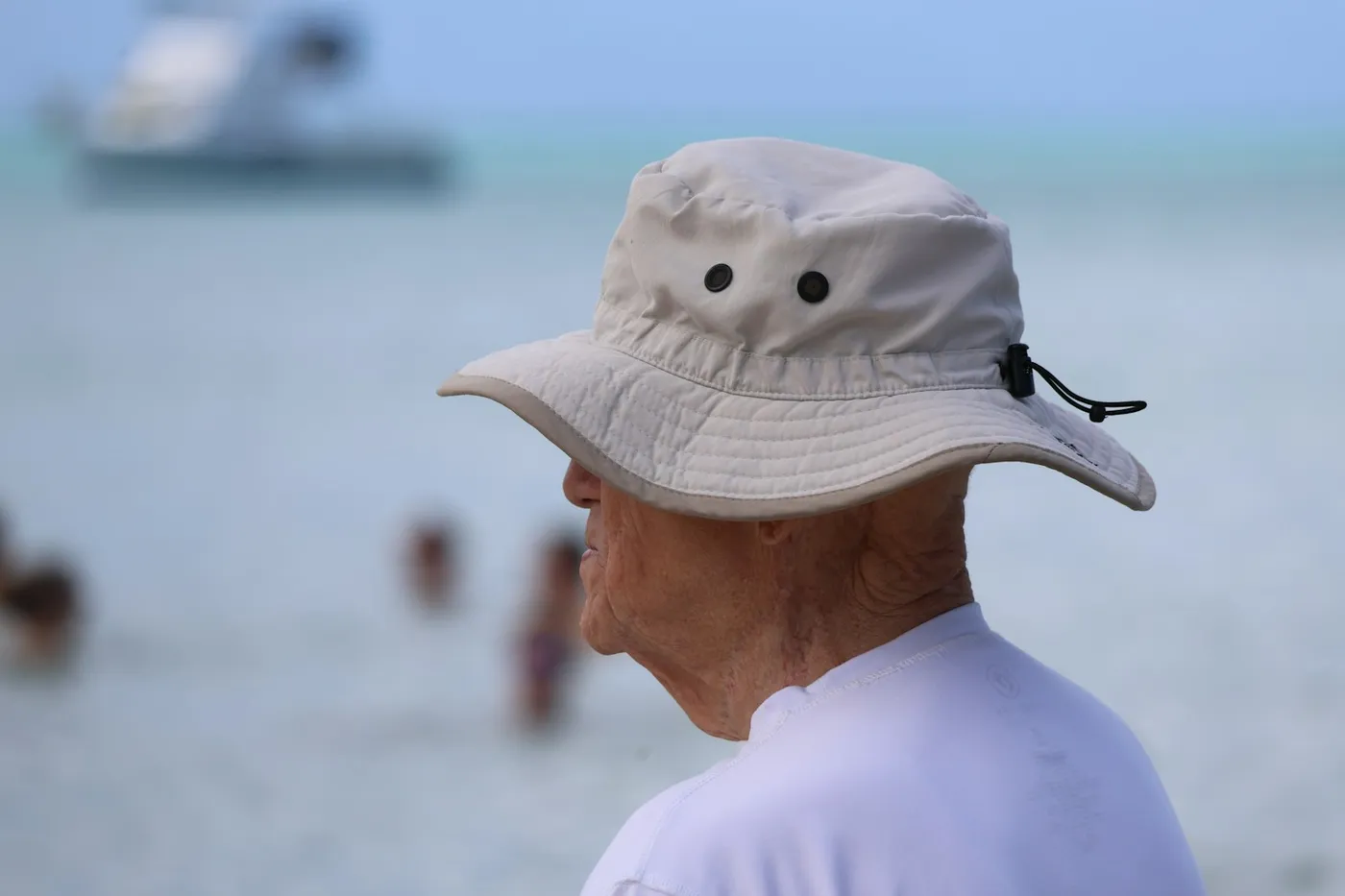 Homem idoso de perfil usando chapéu de aba larga bege e camisa clara de manga, à beira do mar