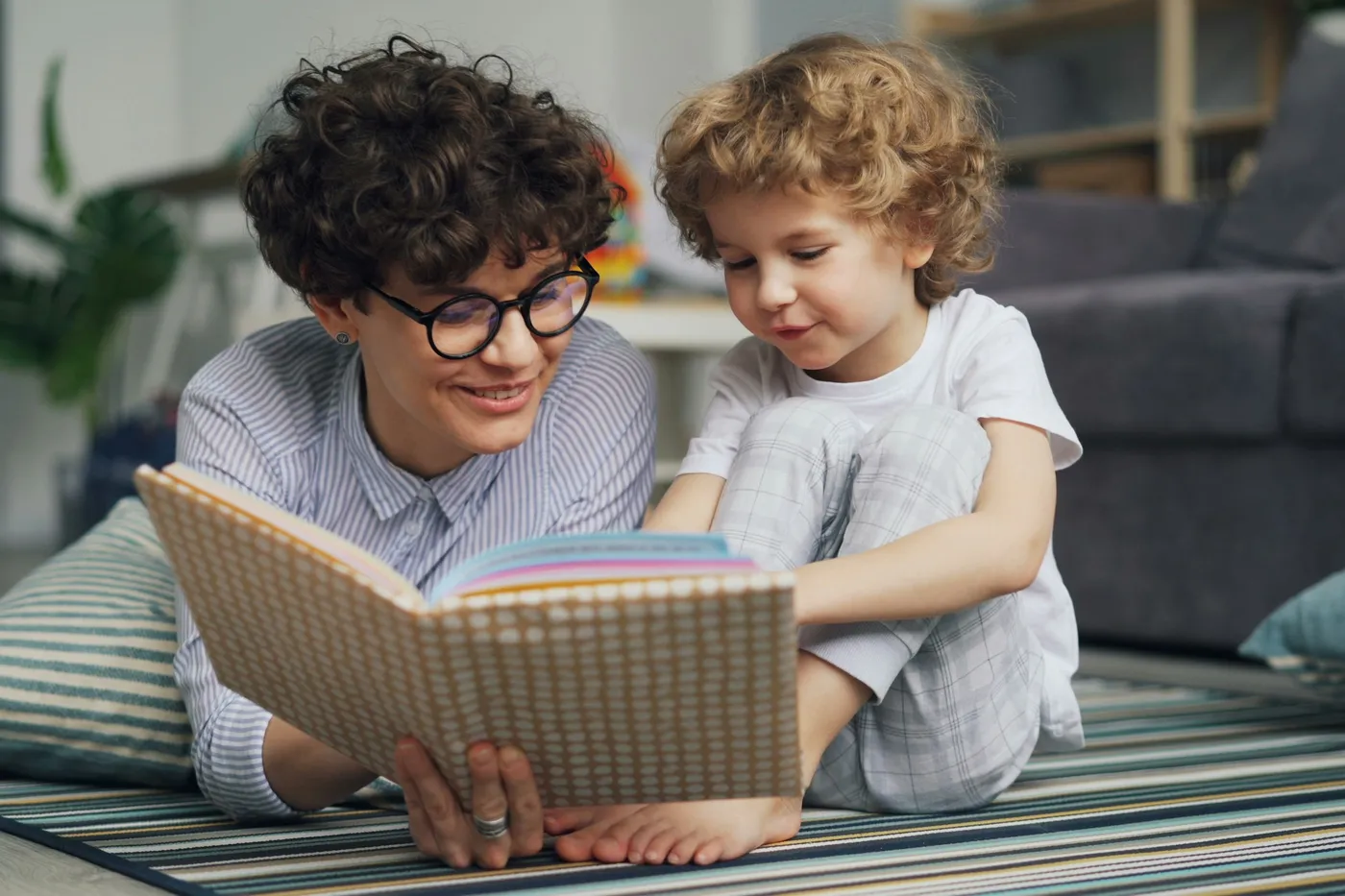 Mulher de óculos e cabelo cacheado deitada num tapete listrado lê um livro de capa estampada com um menino de pijama xadrez sentado ao lado dela