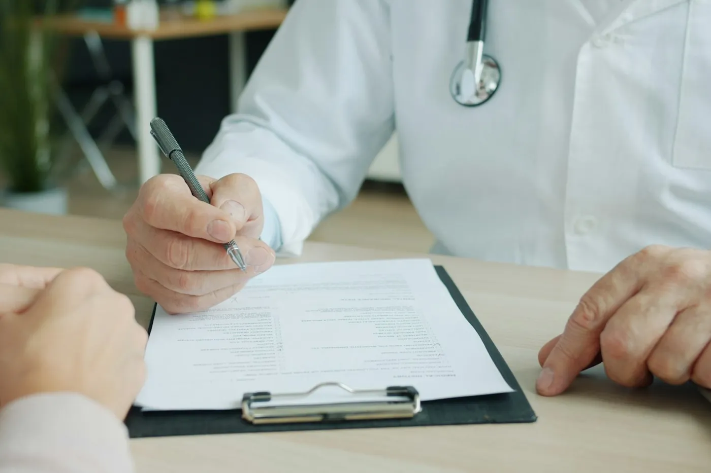 Médico de jaleco branco e estetoscópio preenchendo um formulário numa prancheta sobre a mesa, diante das mãos cruzadas do paciente