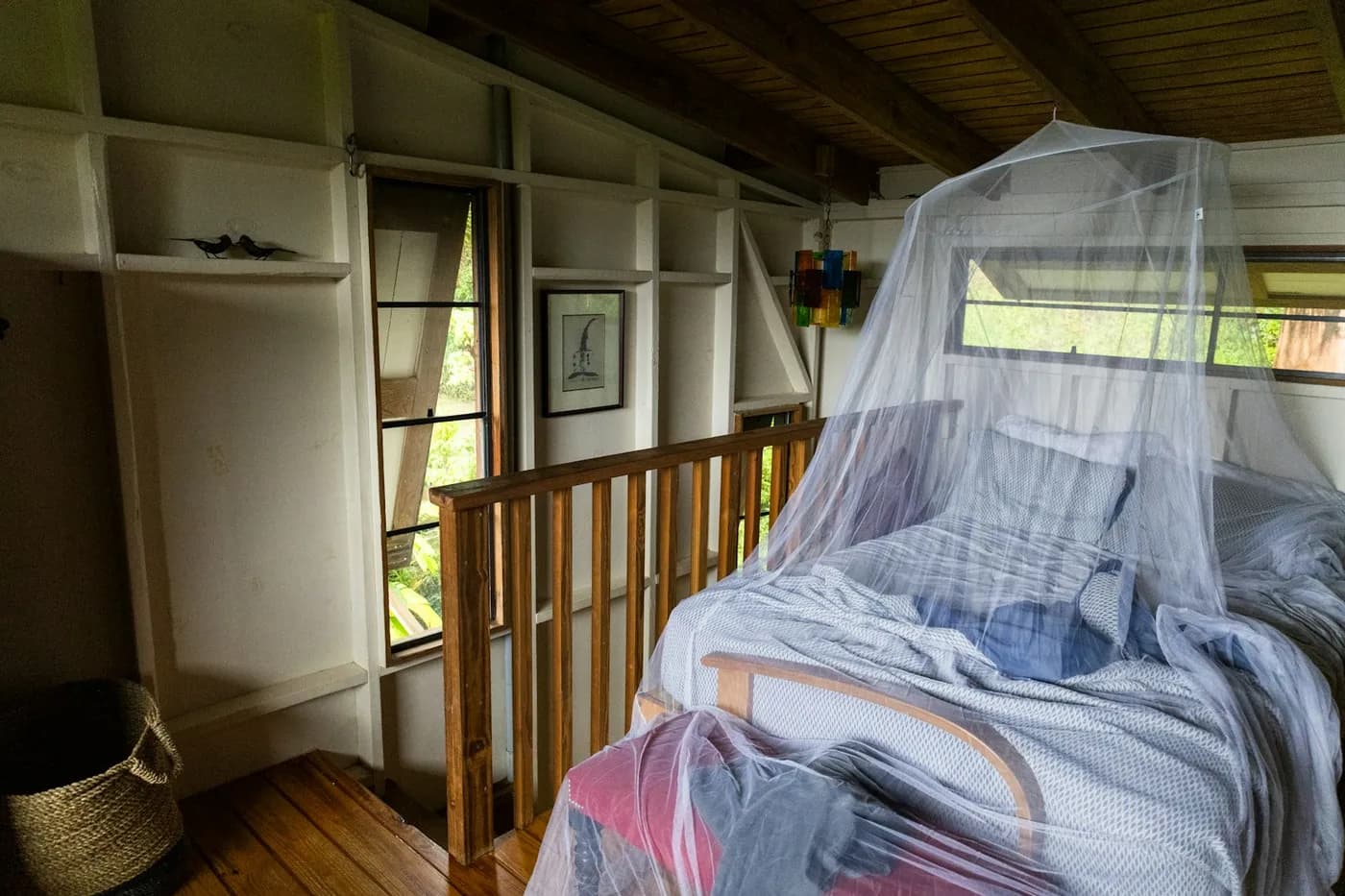 Quarto com paredes de madeira e cama coberta por um mosquiteiro branco, com janela aberta para a vegetação