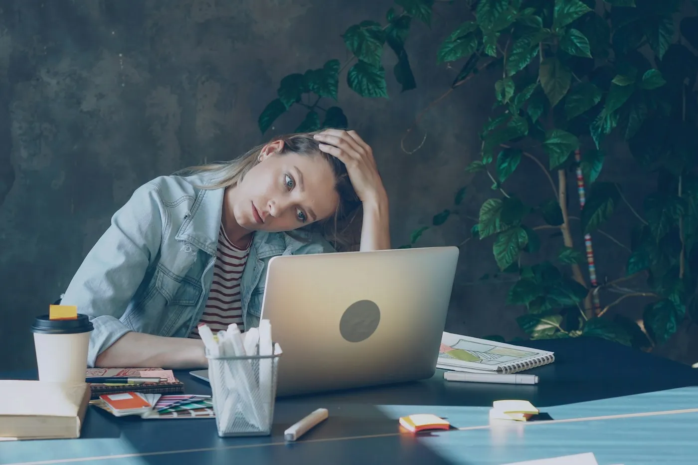 Mulher de jaqueta jeans com a mão na cabeça e expressão cansada diante de um notebook, numa mesa de trabalho com papéis e copo de café