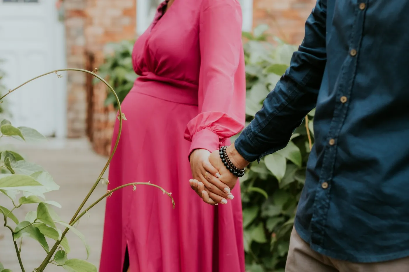 Gestante de vestido longo rosa de mãos dadas com um homem de camisa azul-marinho num jardim com folhagens