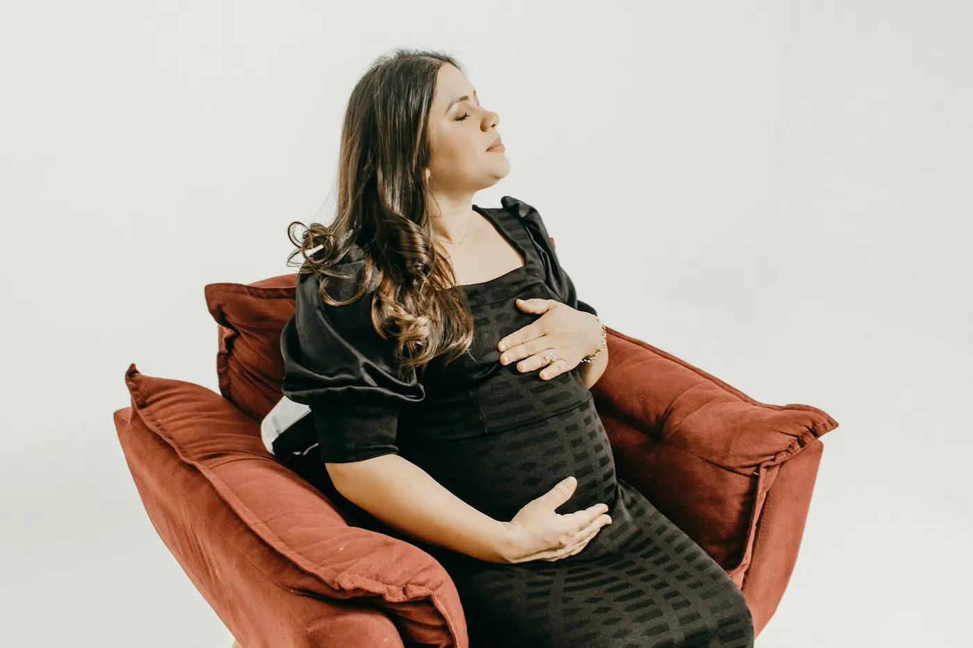 Gestante de vestido preto sentada em uma poltrona vermelha, de olhos fechados e com as mãos sobre a barriga