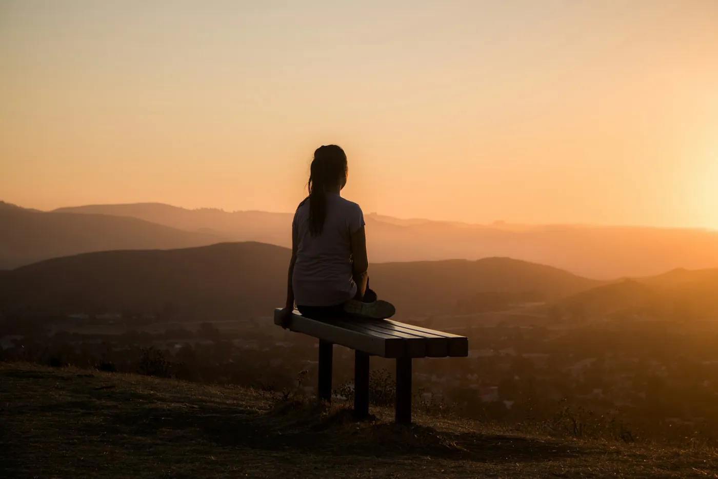 Mulher sentada num banco diante do pôr do sol nas montanhas