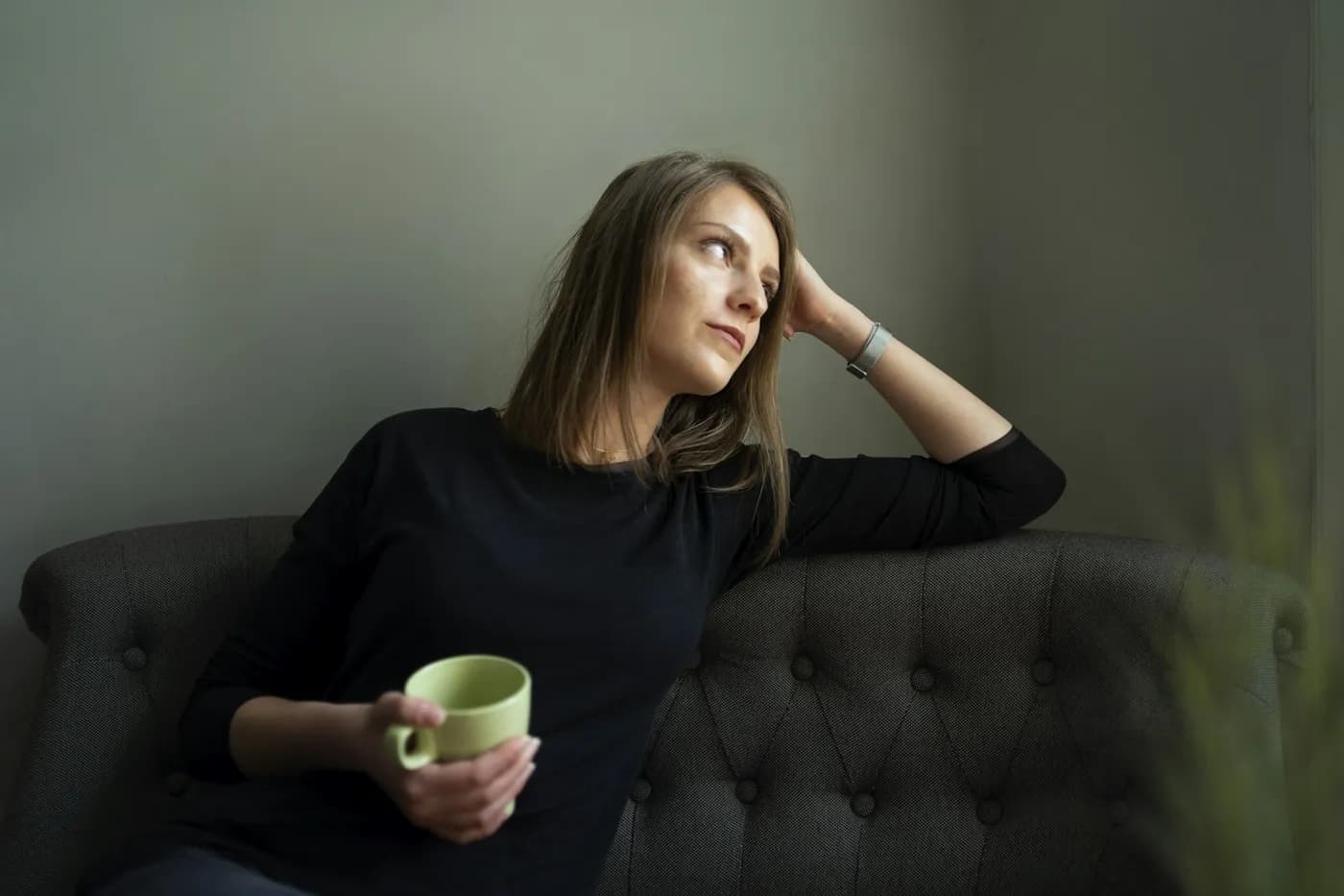 Mulher de blusa preta recostada em um sofá cinza capitonê, segurando uma caneca verde e olhando pensativa para o lado