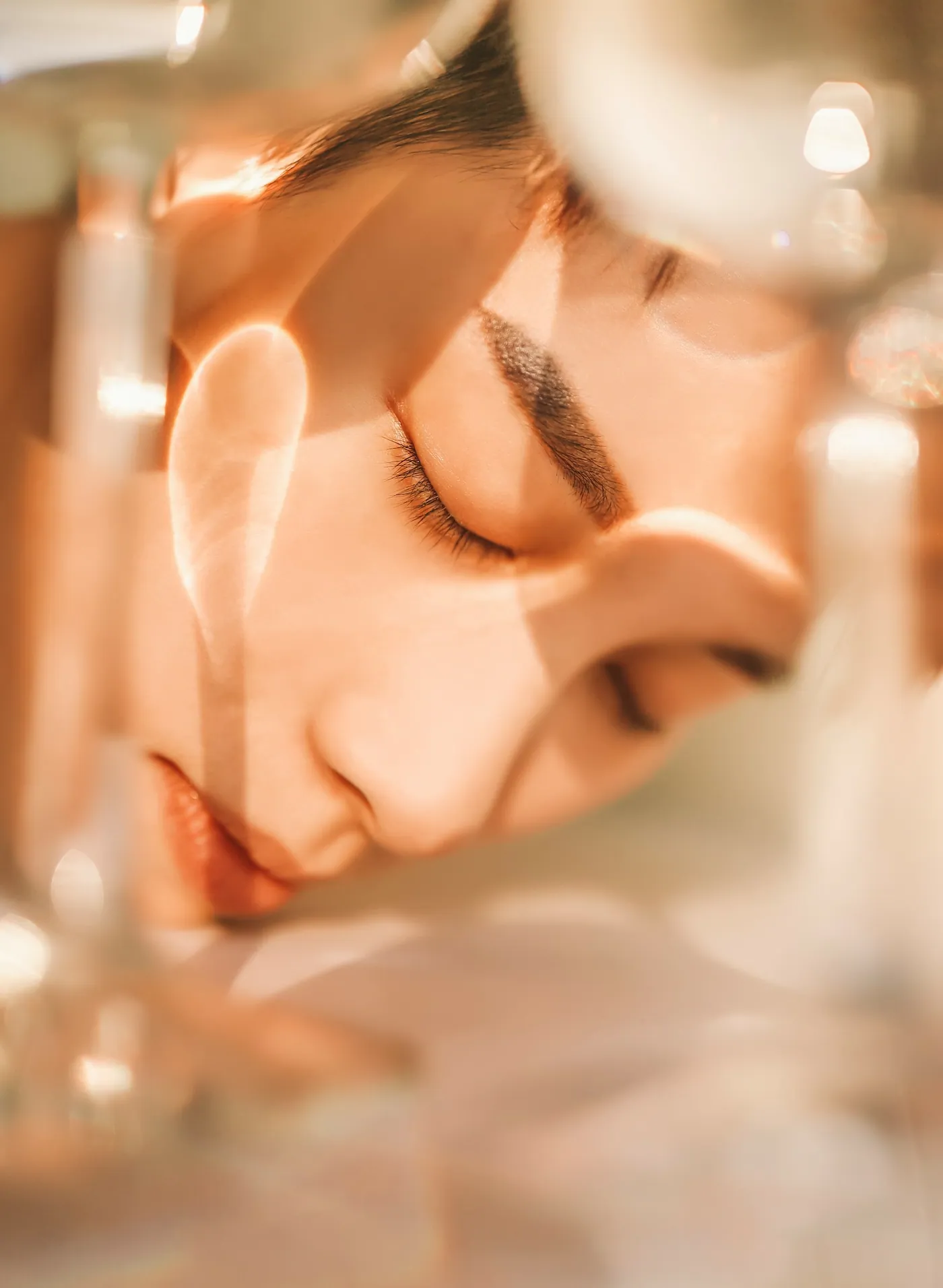 Close no rosto de uma mulher de olhos fechados, com reflexos de luz dourada atravessando objetos de vidro sobre a pele