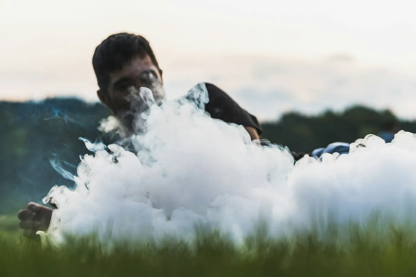 Rapaz deitado na grama ao entardecer, com o rosto parcialmente encoberto por uma nuvem densa de vapor branco