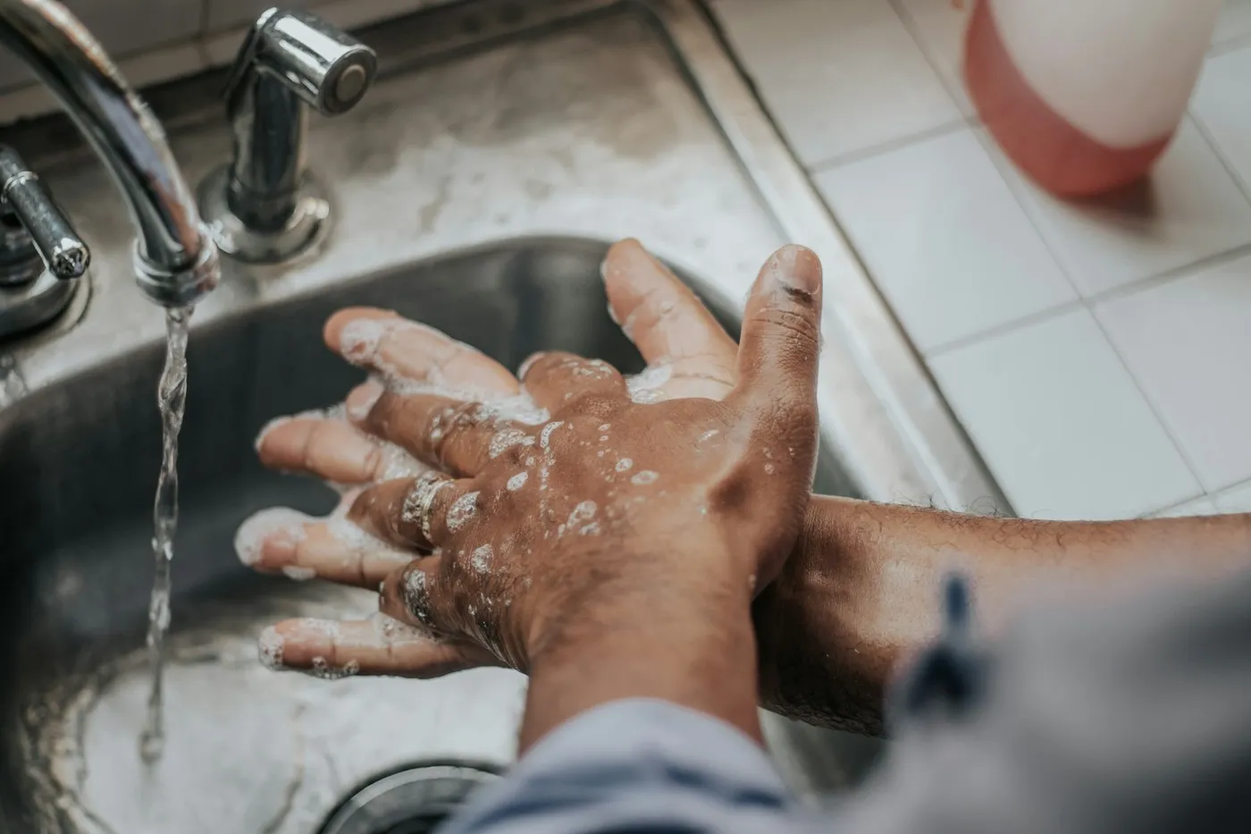 Mãos ensaboadas sendo lavadas sob a torneira de uma pia de inox, com um frasco de sabonete ao fundo