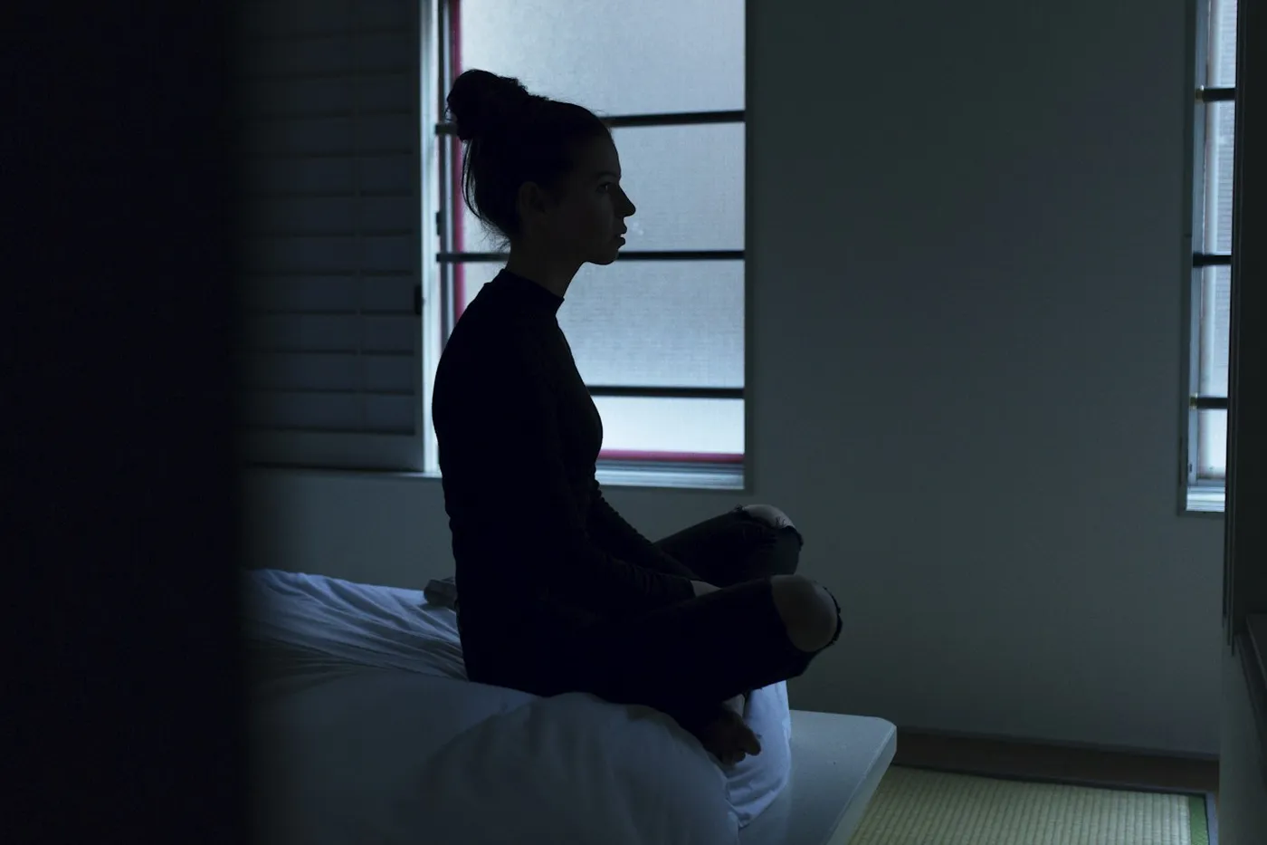 Mulher de blusa escura e cabelo preso sentada de pernas cruzadas sobre a cama, de perfil, olhando para a janela num quarto em penumbra