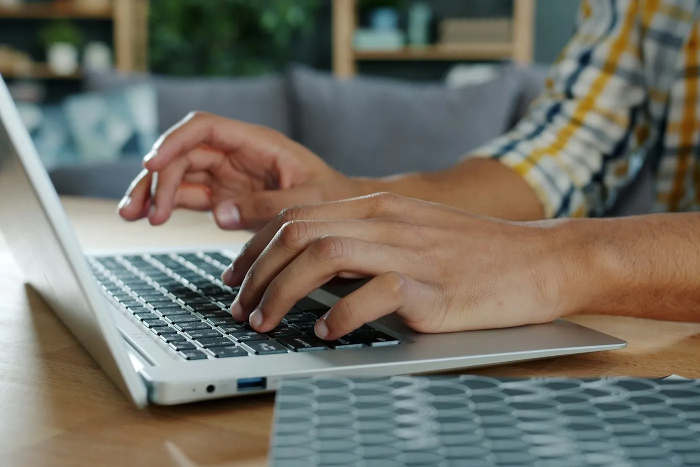 Mãos de uma pessoa digitando em um notebook prateado apoiado em uma mesa de madeira, em uma sala com sofá ao fundo