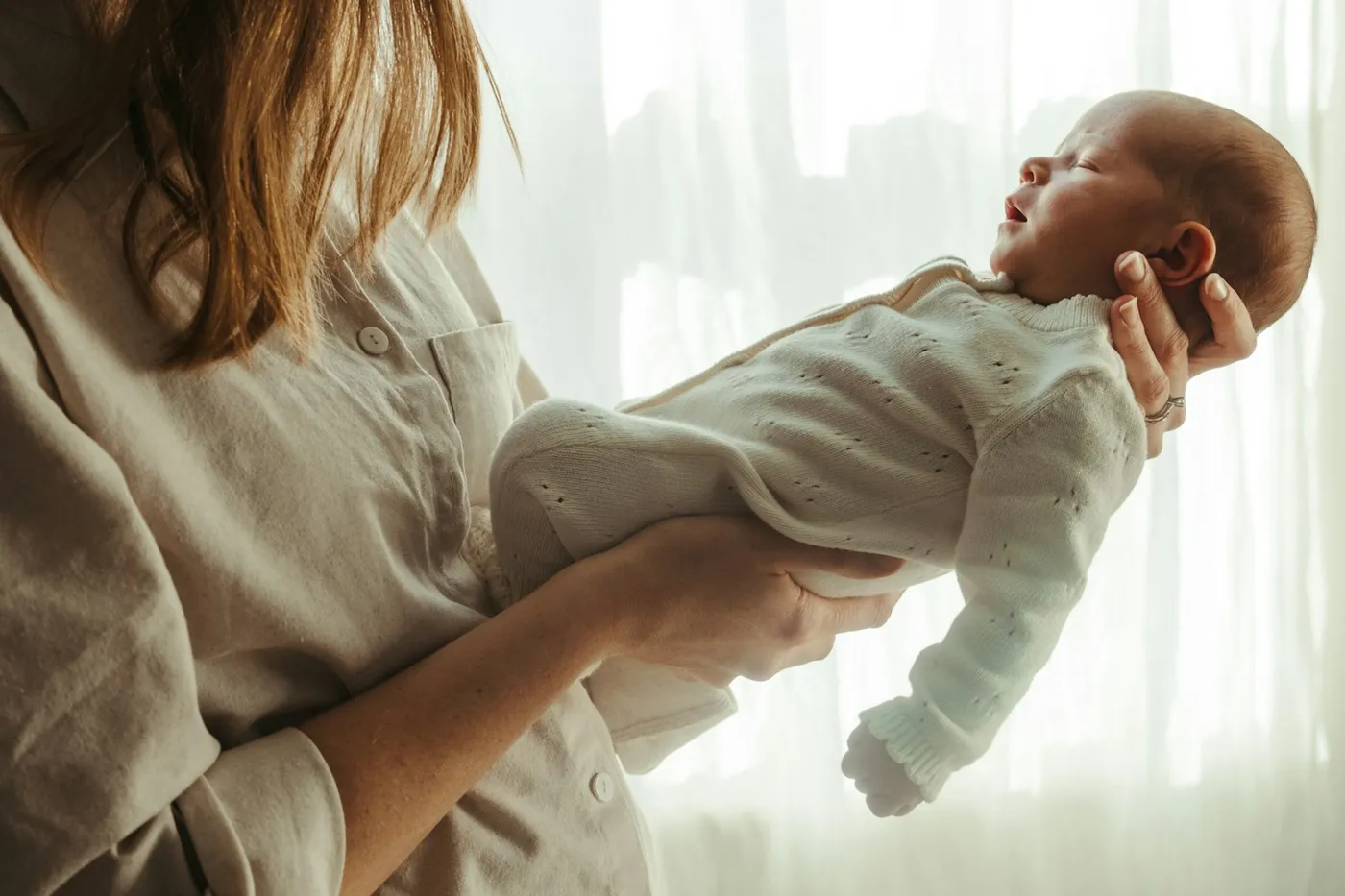 Mãe de camisa bege segurando nos braços um recém-nascido adormecido, de macacão branco de tricô, diante de uma cortina iluminada