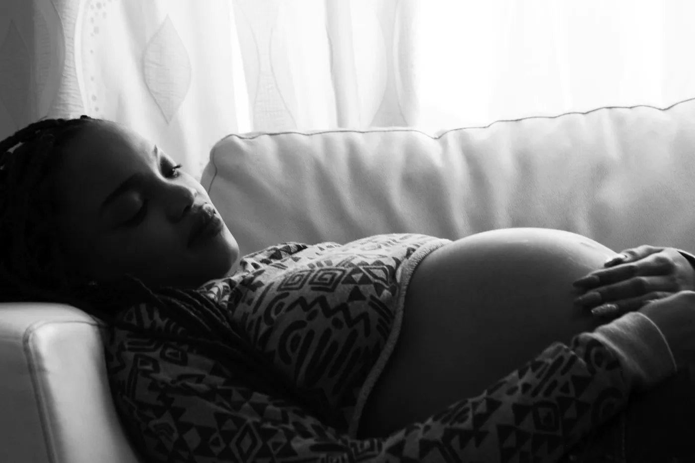Foto em preto e branco de uma gestante deitada em um sofá claro, de olhos fechados e com as mãos sobre a barriga
