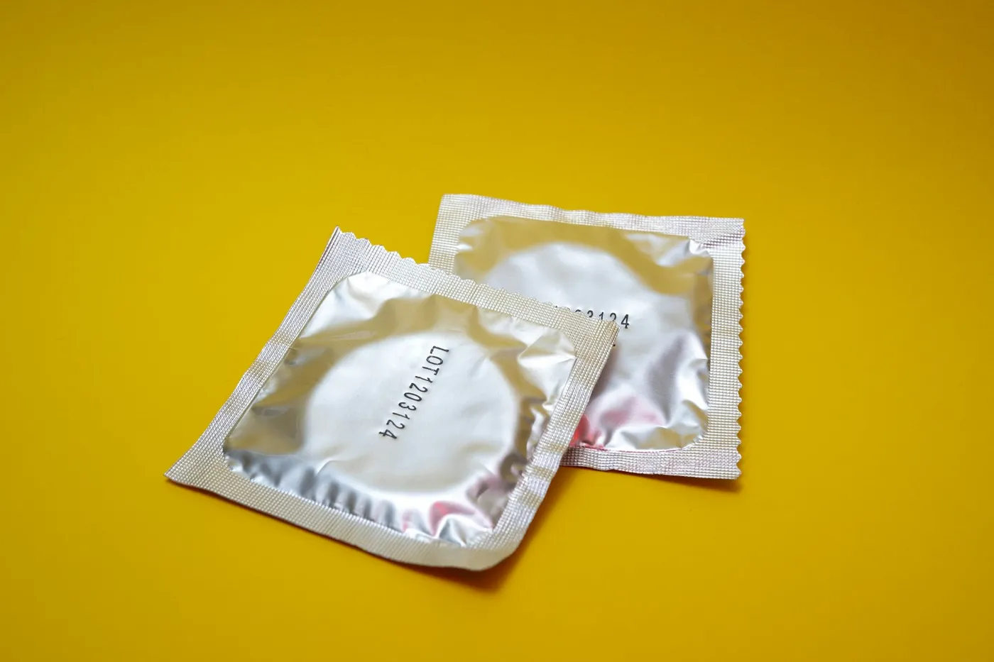 Duas embalagens fechadas de preservativo, prateadas, sobre um fundo amarelo