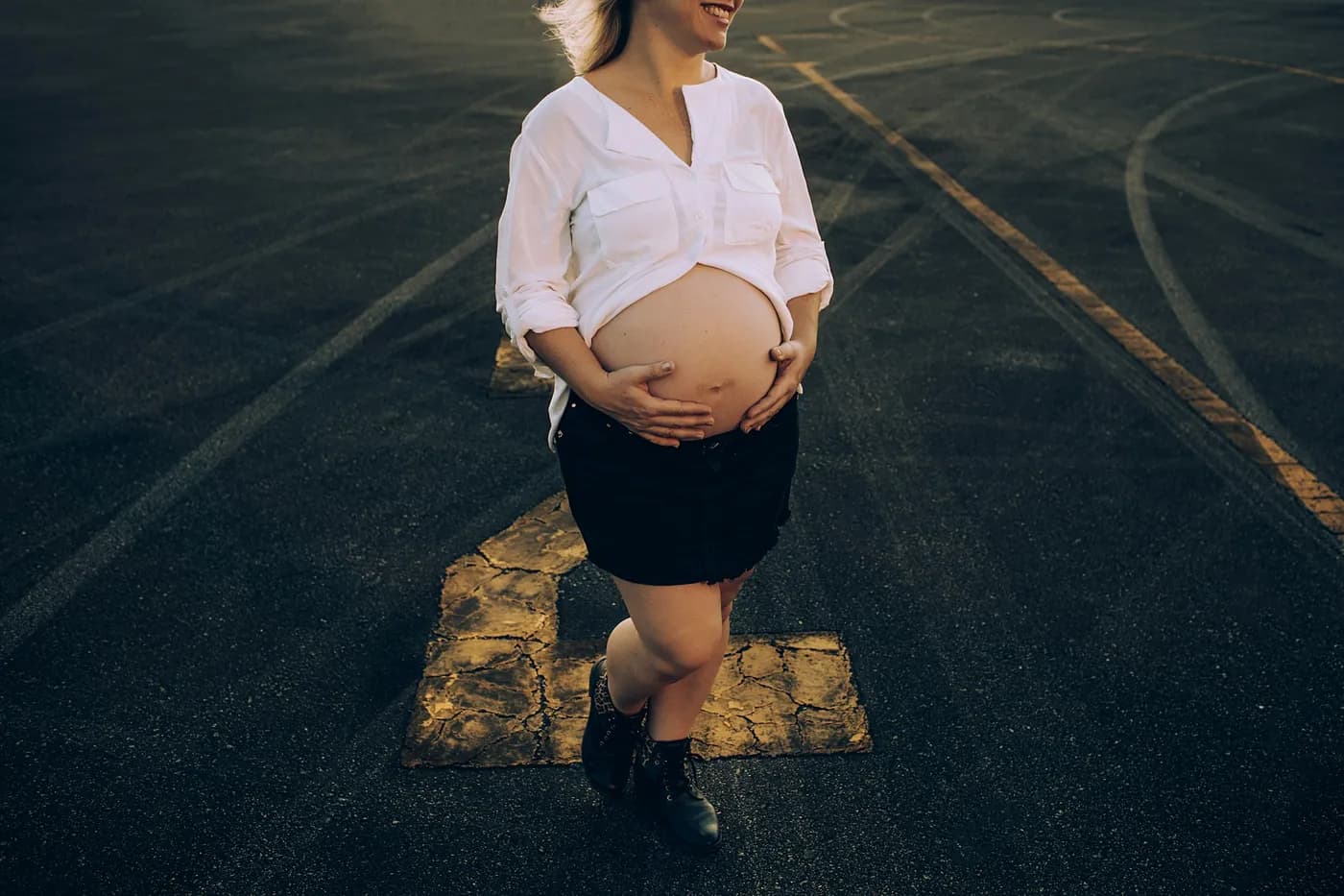 Gestante de camisa branca e saia preta caminhando sobre o asfalto ao fim da tarde, com as mãos apoiadas na barriga