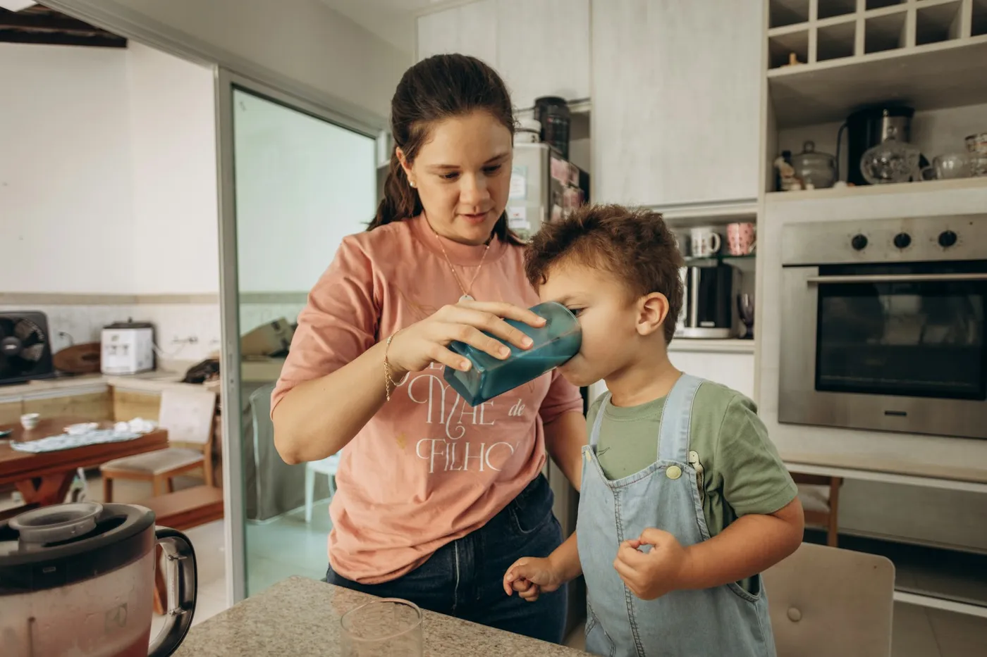 Mãe de camiseta rosa ajudando o filho pequeno, de jardineira jeans, a beber de um copo azul na cozinha de casa