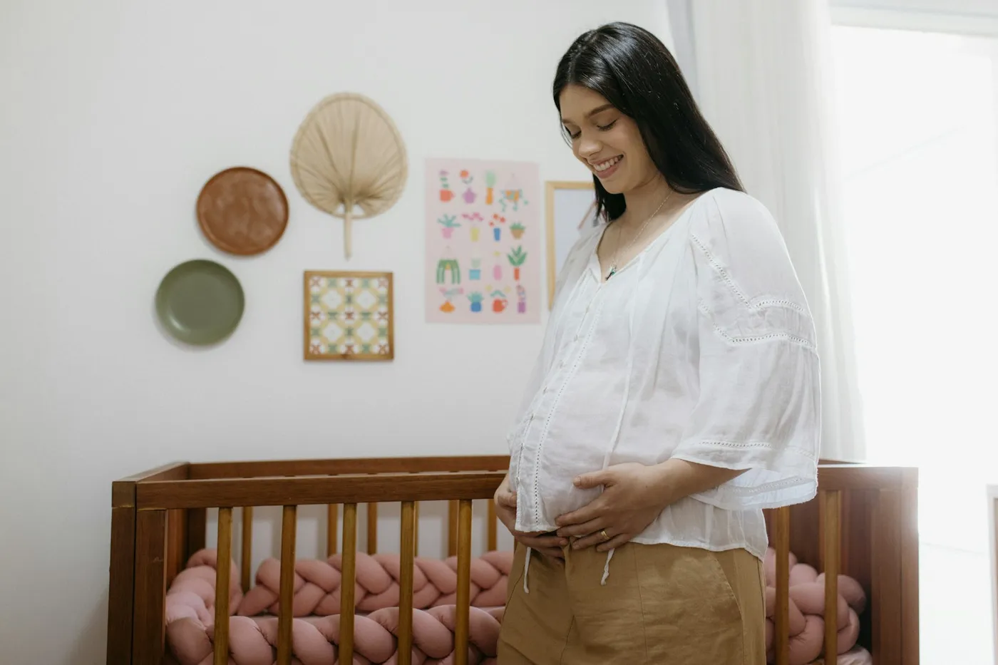 Gestante sorridente de blusa branca, com as mãos na barriga, em pé diante de um berço de madeira num quarto de bebê