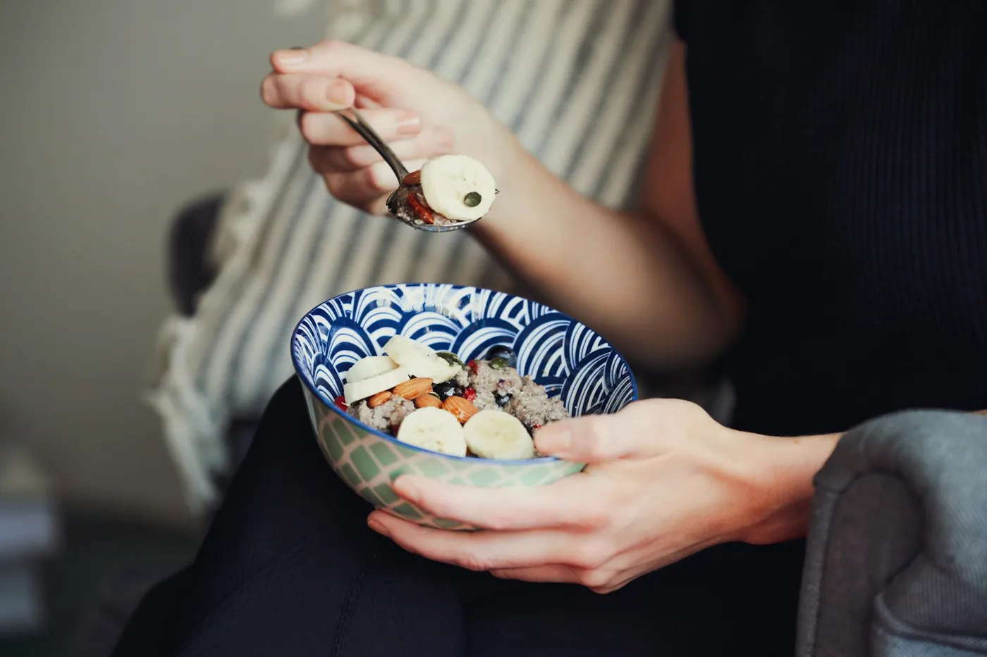 Mãos de uma mulher de blusa preta segurando uma tigela de louça com estampa azul de ondas, com mingau de grãos, rodelas de banana, amêndoas e frutas vermelhas, e levando uma colherada à boca