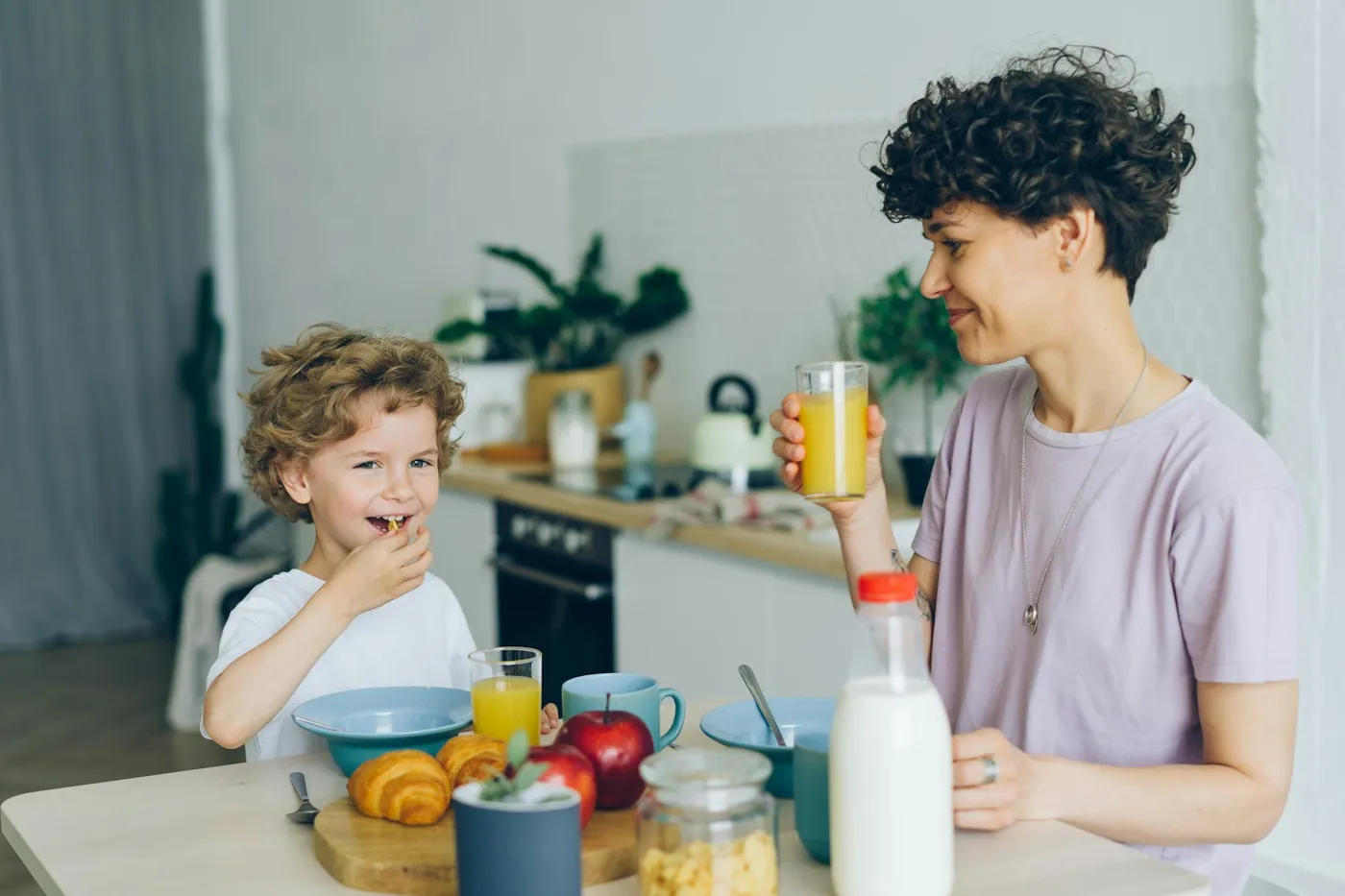 Menino de cabelo cacheado sentado à mesa do café da manhã comendo cereal, ao lado da mãe que bebe suco de laranja