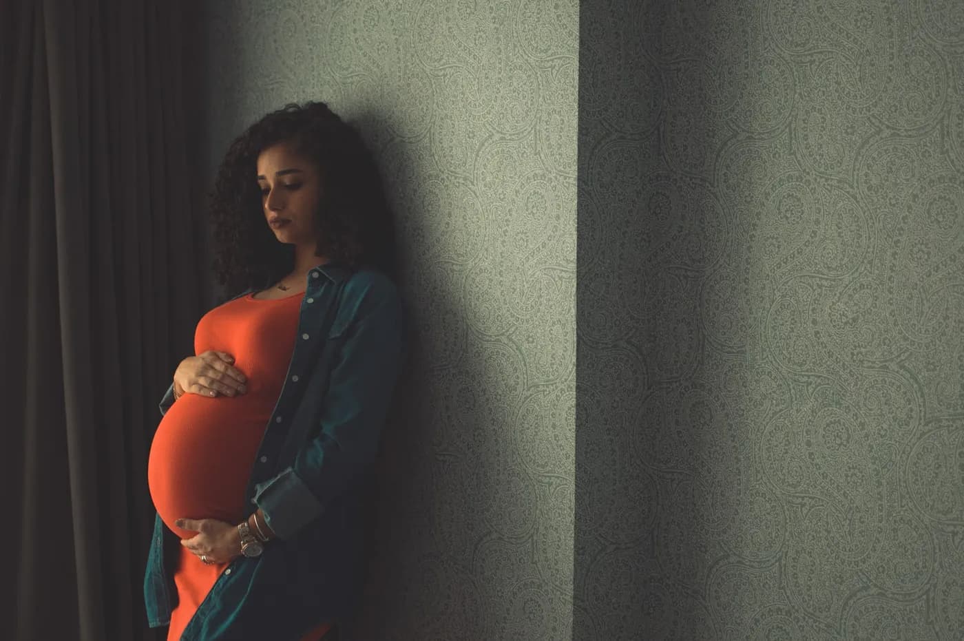 Gestante de blusa laranja e jaqueta jeans encostada em uma parede, com as mãos sobre a barriga