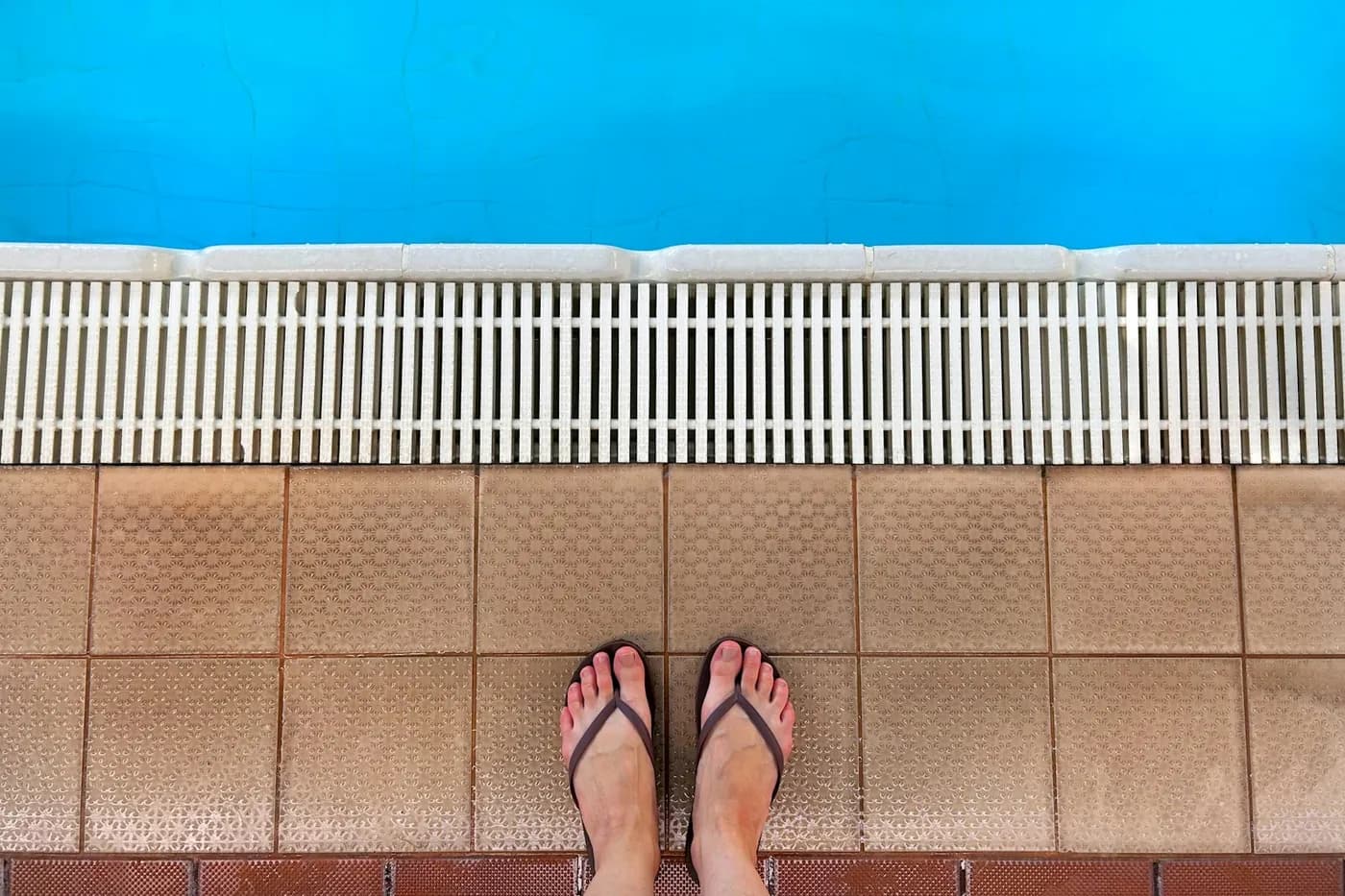 Pés de chinelo de dedo marrom sobre o piso de cerâmica da borda de uma piscina azul, vistos de cima
