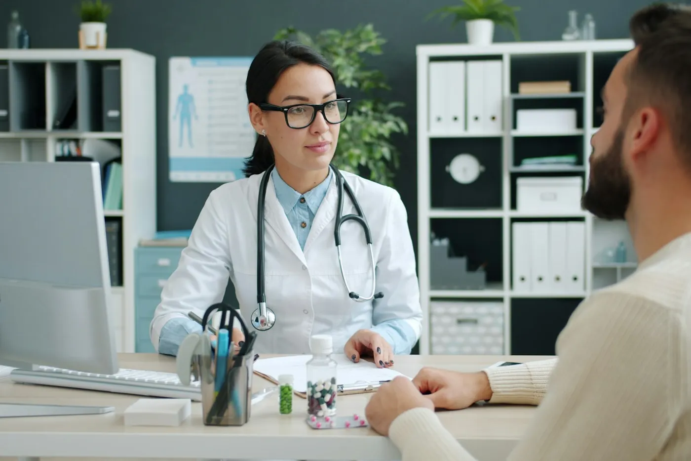 Médica de óculos e jaleco com estetoscópio conversando com um paciente de suéter claro em um consultório com estantes