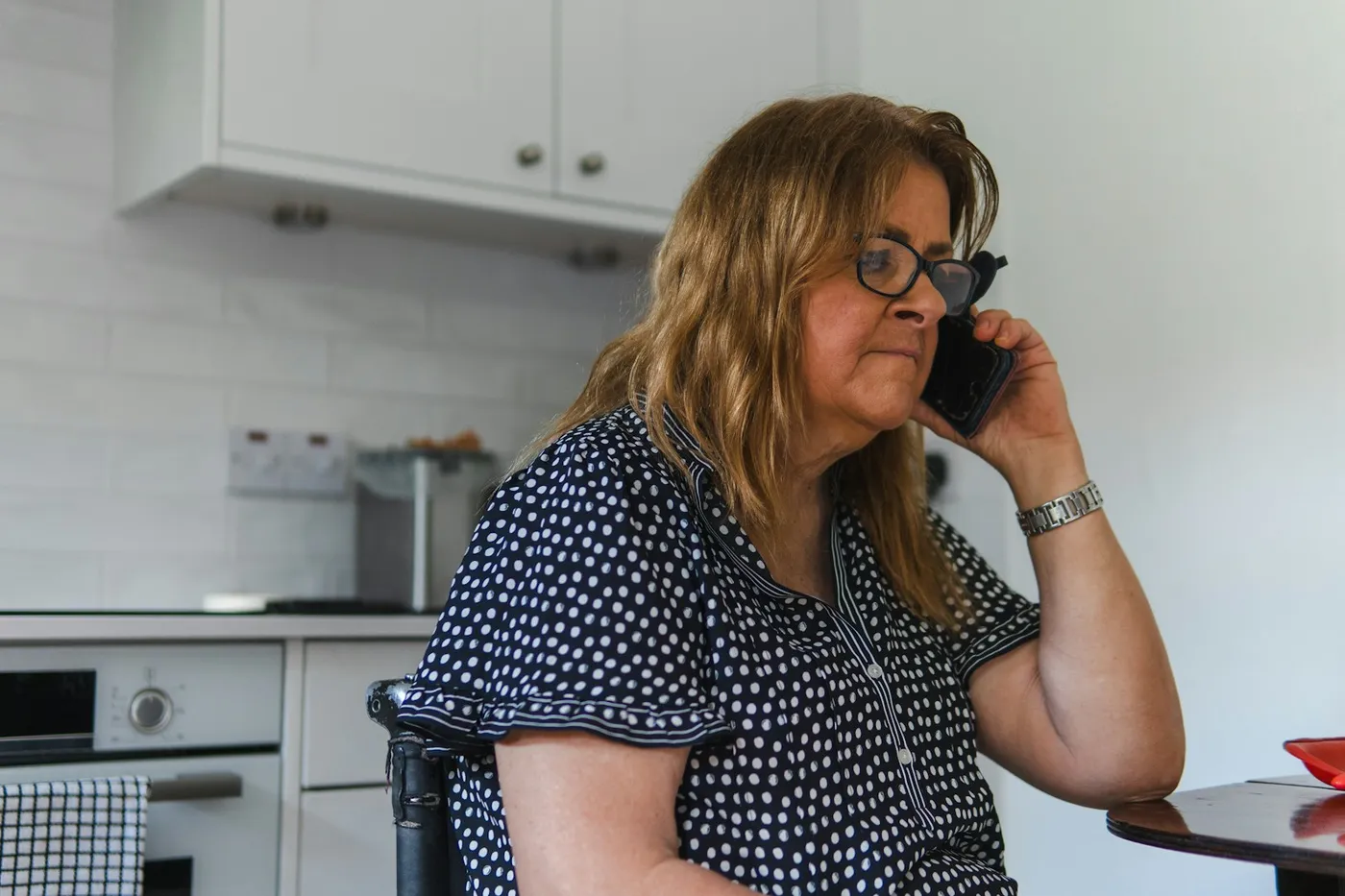 Mulher de óculos e blusa azul-marinho de bolinhas sentada na cozinha, falando ao celular com expressão séria