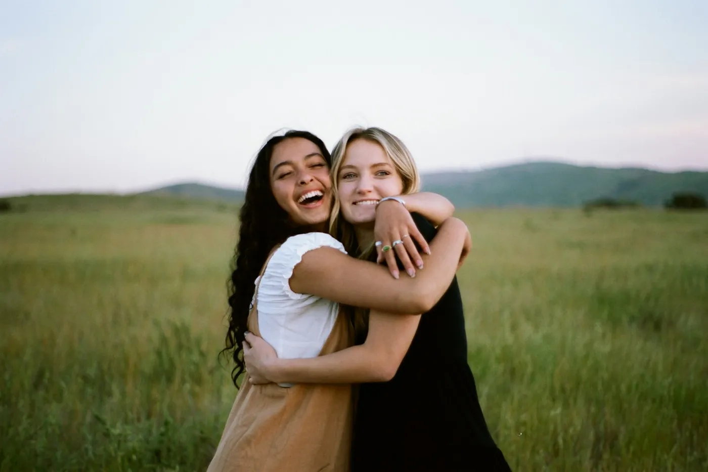 Duas amigas sorrindo e se abraçando em um campo de grama alta, com morros ao fundo
