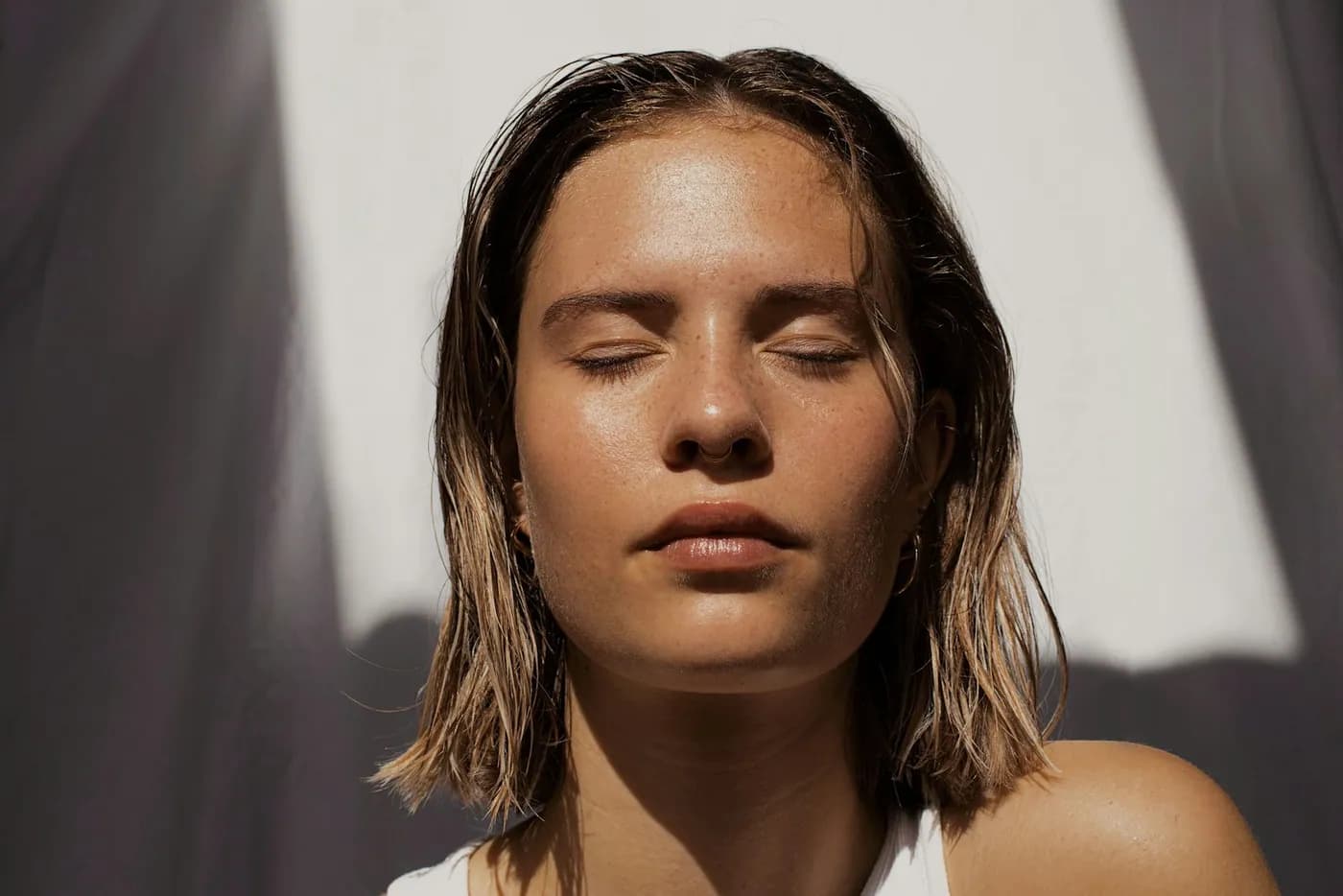 Rosto de mulher com os olhos fechados sob luz forte, cabelo molhado e pele sem maquiagem