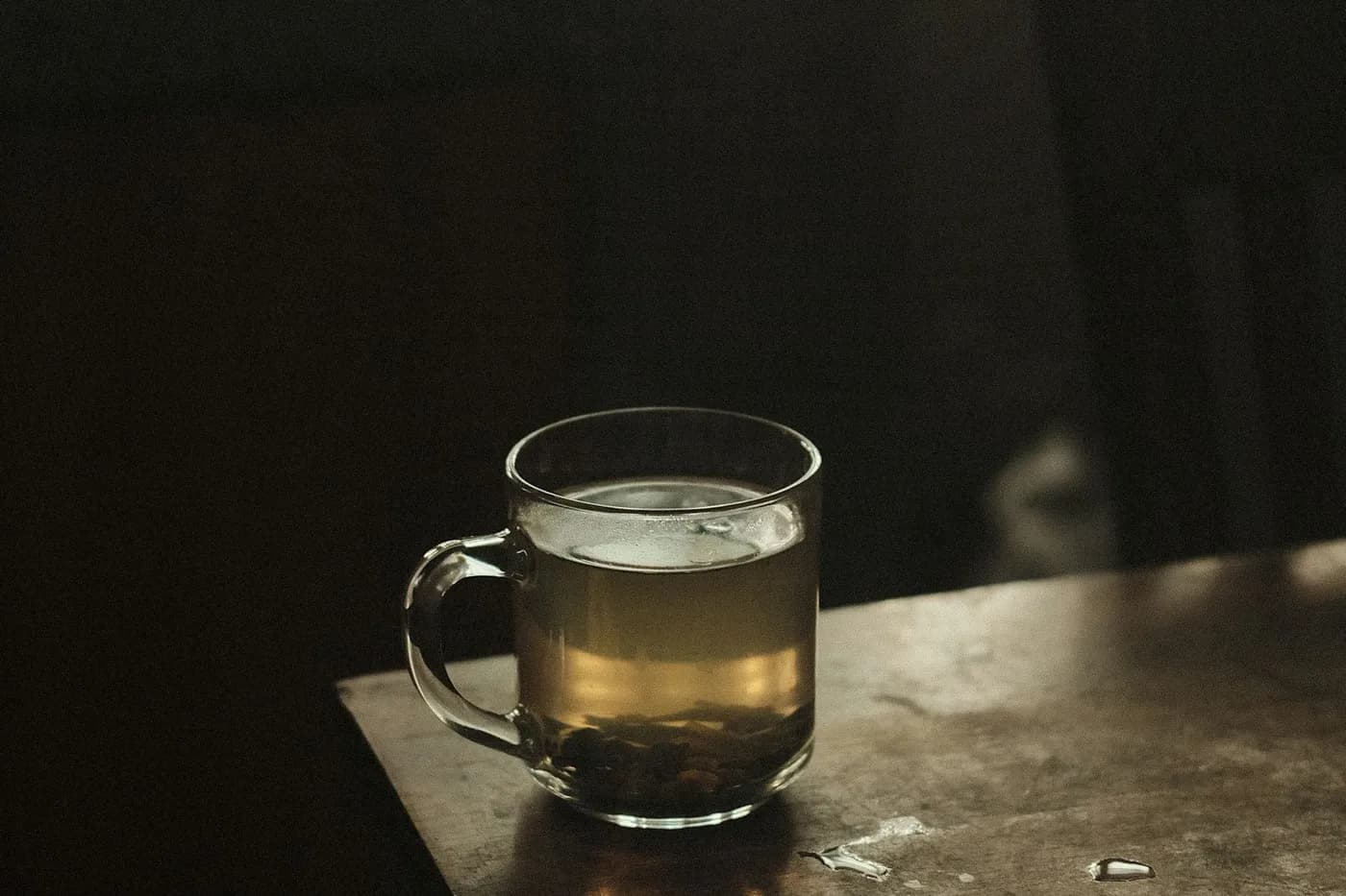Caneca de vidro com chá de ervas amarelado e folhas no fundo, sobre uma mesa de madeira gasta, num ambiente escuro iluminado por luz lateral