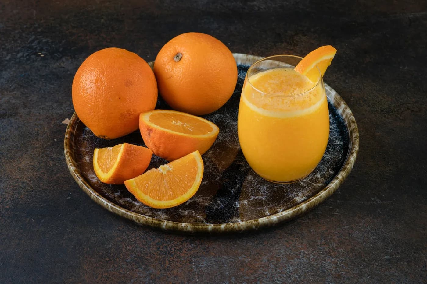Laranjas inteiras e cortadas em gomos ao lado de um copo de suco de laranja sobre uma bandeja escura