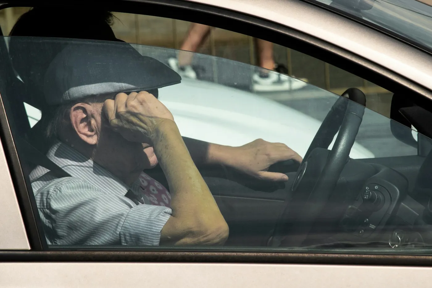 Homem idoso de boina e camisa listrada ao volante de um carro, apoiando a cabeça na mão sobre os olhos, visto pela janela do motorista