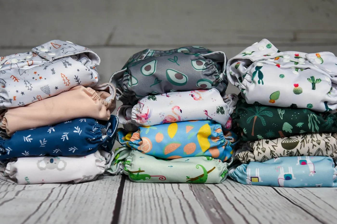 Três pilhas de fraldas de pano reutilizáveis com estampas de abacate, flamingo, cacto e folhas sobre um piso de madeira