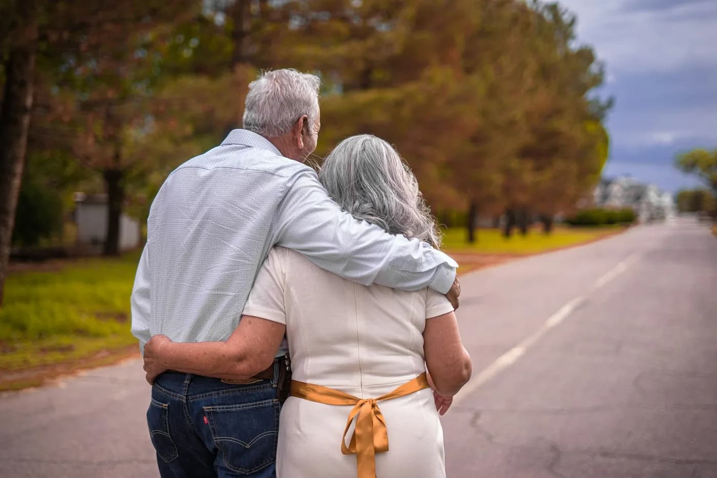 Casal de idosos de cabelos grisalhos caminhando abraçado por uma rua arborizada, vistos de costas