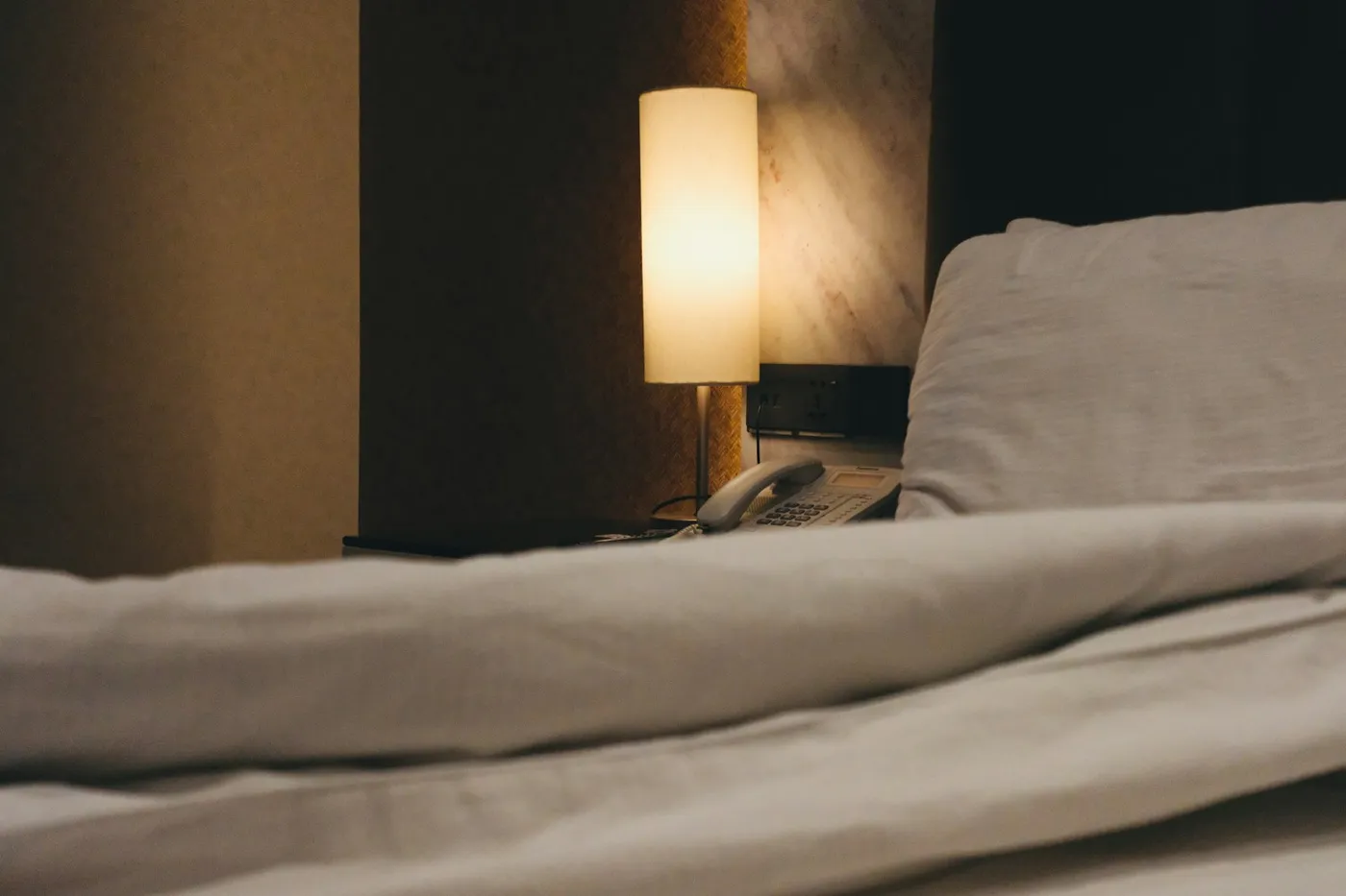 Quarto escuro com abajur aceso sobre a mesa de cabeceira, ao lado de uma cama com edredom branco