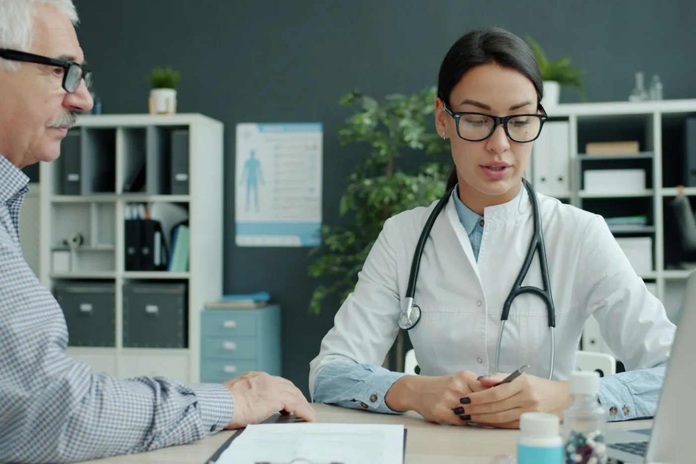 Médica de jaleco e estetoscópio conversando com um homem idoso de óculos sentado à mesa do consultório