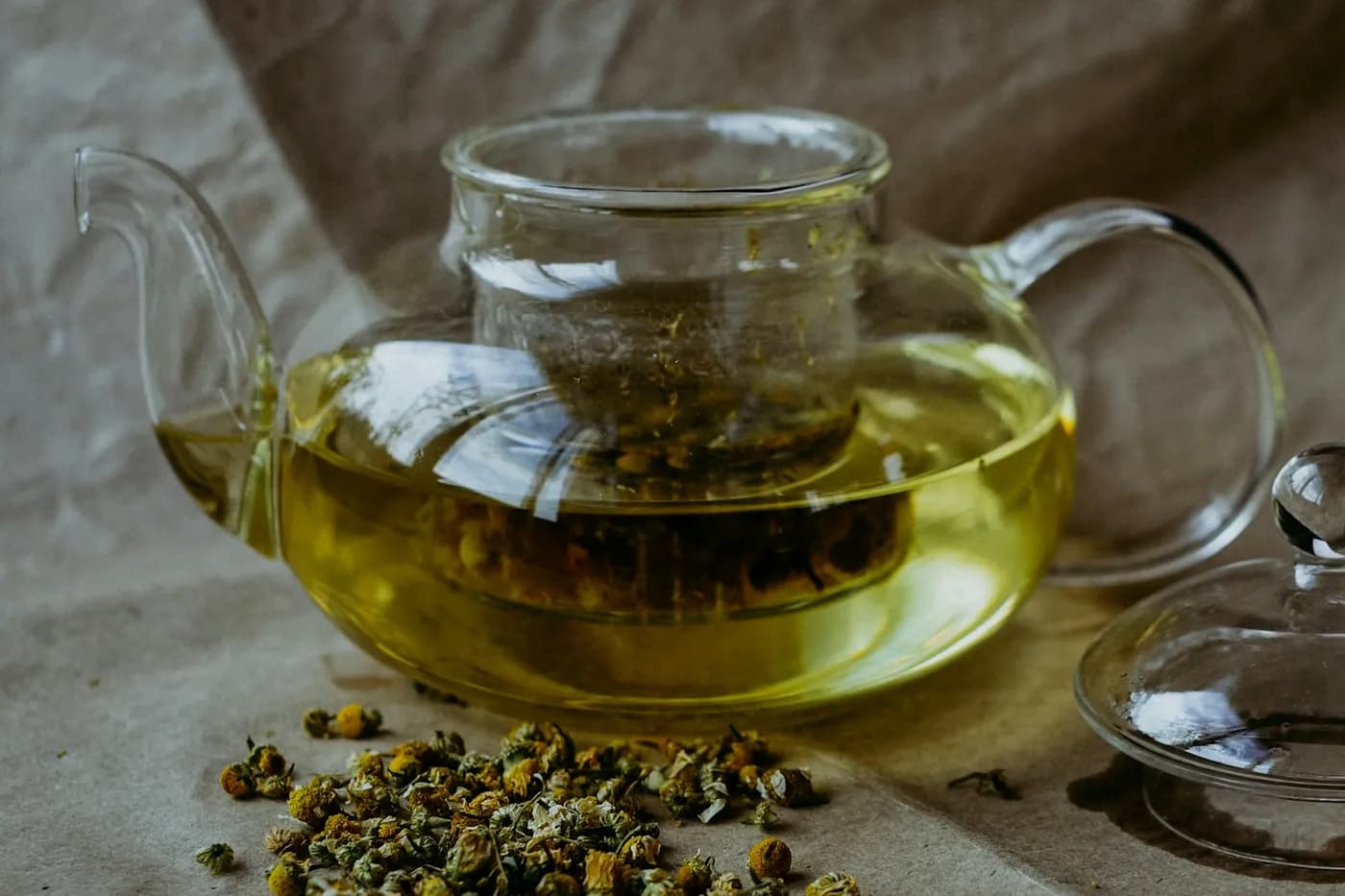 Bule de vidro com infusão amarelo-esverdeada e flores secas de camomila no filtro, com um punhado de flores secas espalhado sobre papel pardo e a tampa de vidro ao lado
