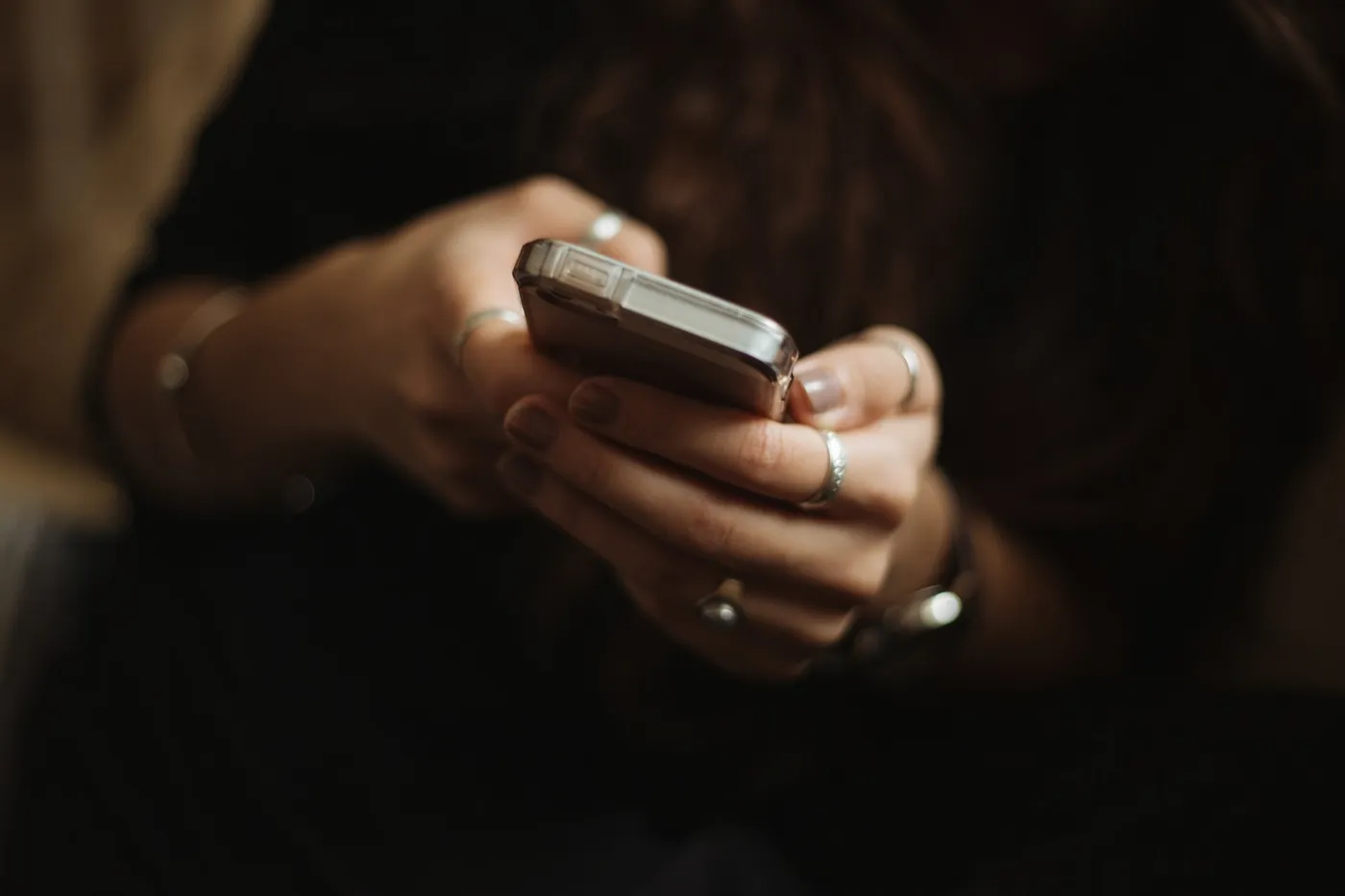 Mãos de uma pessoa com anéis prateados segurando um celular em um ambiente com pouca luz