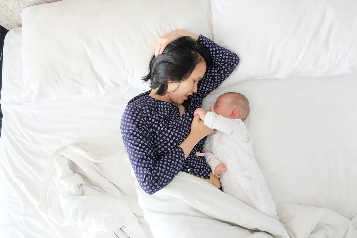 Mãe de blusa azul-marinho com estampa miúda deitada de lado numa cama de lençóis brancos, de frente para o bebê de macacão branco, que segura a mão dela