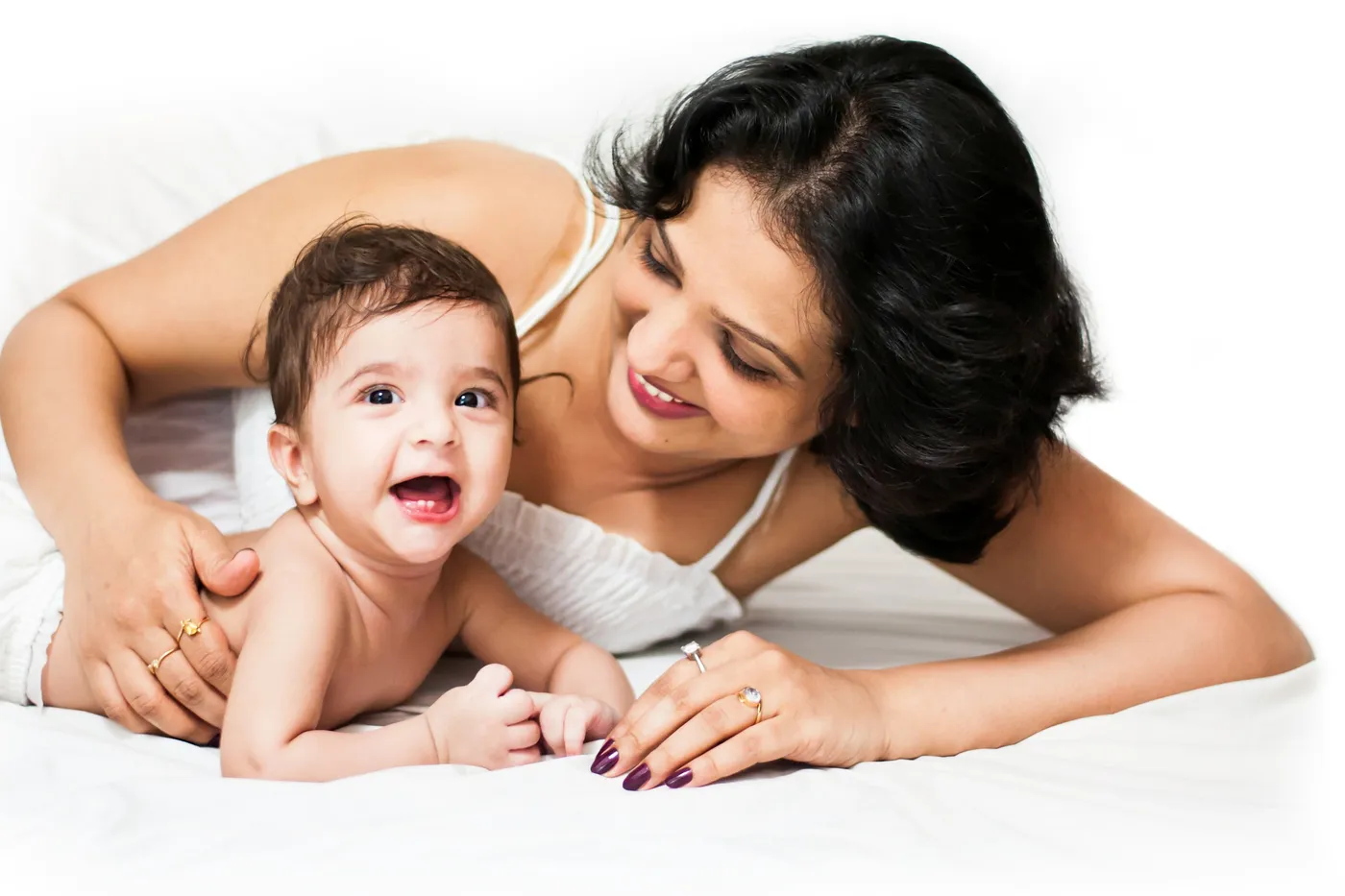 Mulher de cabelo preto e blusa branca deitada de bruços numa cama branca olha sorrindo para um bebê que ri, deitado de barriga para baixo ao lado dela