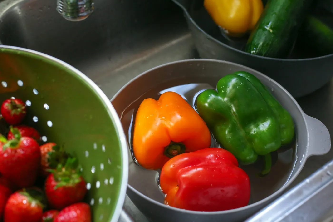 Pimentões laranja, vermelho e verde de molho em uma bacia cinza com água dentro da pia, ao lado de um escorredor verde com morangos