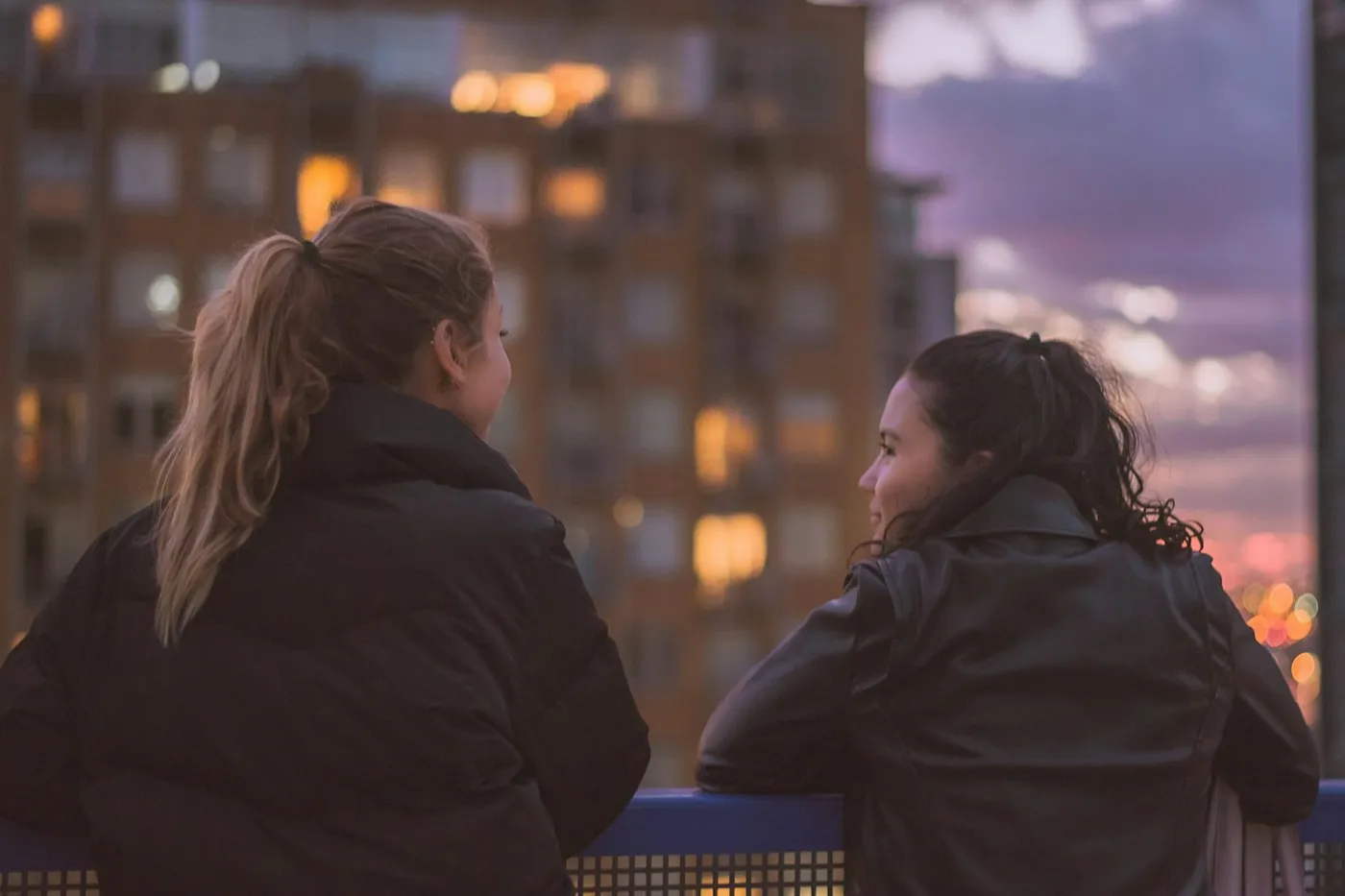 Duas mulheres de casaco escuro conversando lado a lado, apoiadas numa grade, com prédios iluminados ao entardecer
