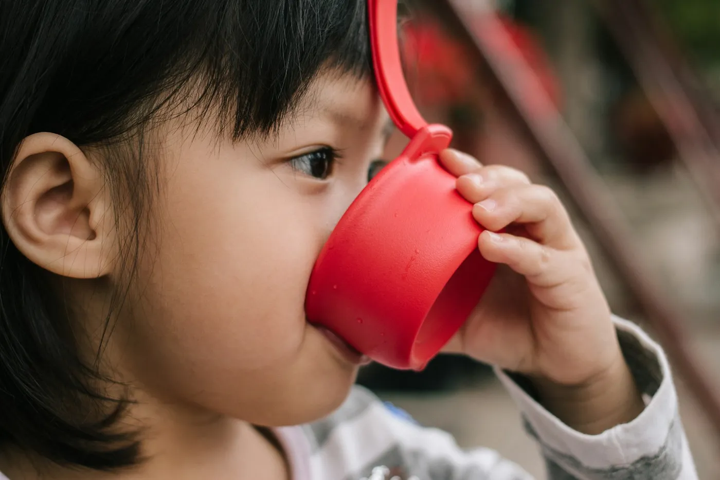 Menina pequena de franja bebendo em uma caneca vermelha de plástico, segurando a alça com a mão