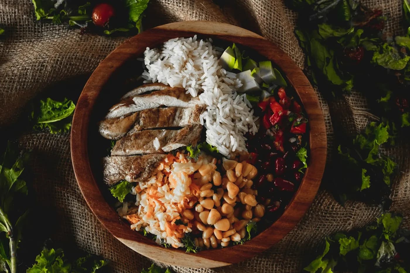 Tigela de madeira vista de cima com arroz branco, frango grelhado fatiado, grão-de-bico, feijão e salada sobre tecido de juta