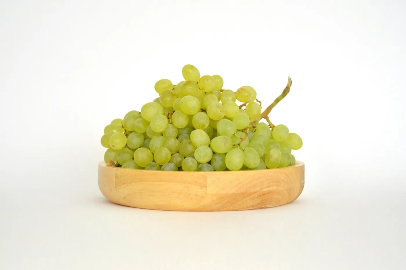 Cacho de uvas verdes sobre um prato raso de madeira clara, em fundo branco