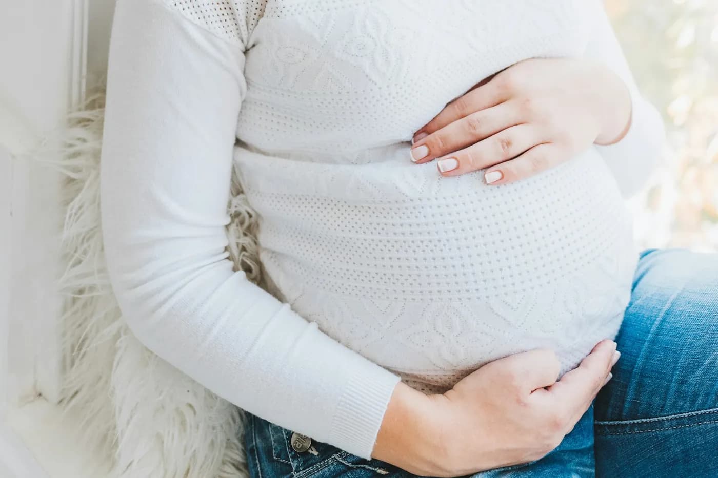 Gestante de suéter branco de tricô e calça jeans sentada perto da janela, com as duas mãos apoiadas na barriga
