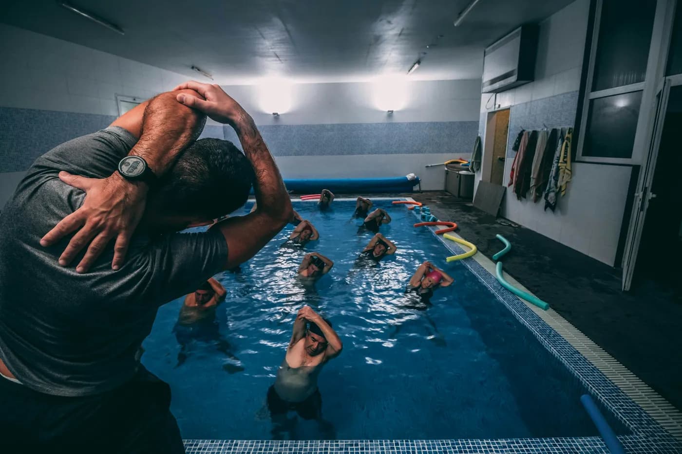Professor de camiseta cinza alongando os braços atrás da cabeça à beira de uma piscina coberta, onde alunos de touca repetem o movimento dentro da água