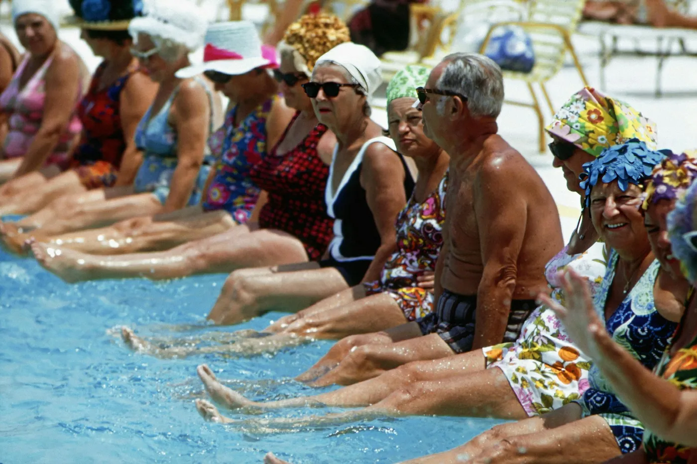 Grupo de idosos sentados na borda de uma piscina, de chapéu e óculos escuros, batendo os pés na água durante uma aula de exercício