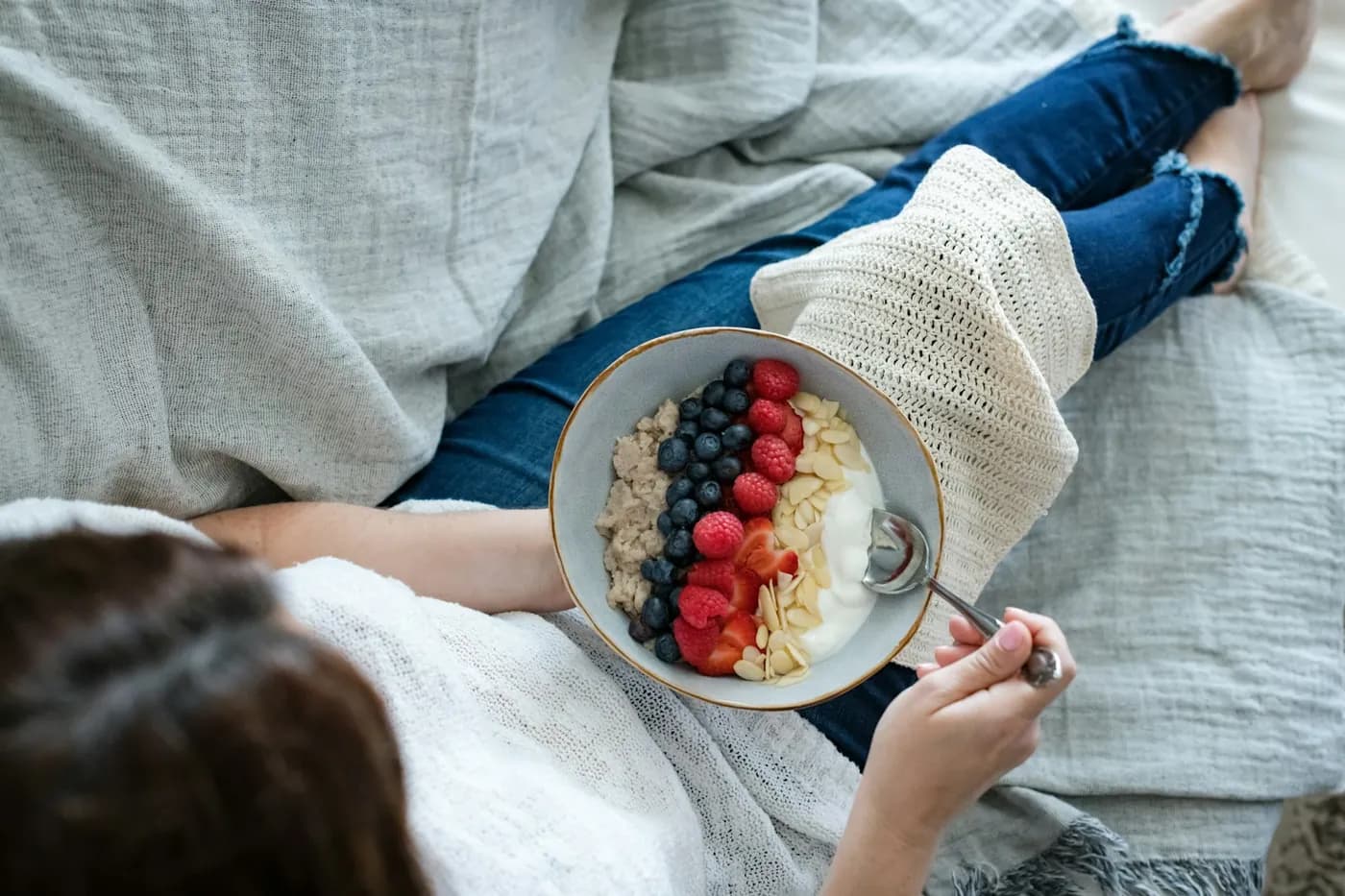 Mulher sentada no sofá com uma tigela de aveia, iogurte, frutas vermelhas e amêndoas