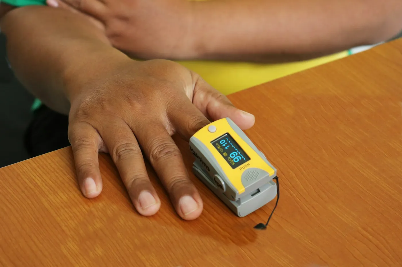 Mão apoiada numa mesa de madeira com um oxímetro amarelo no dedo mostrando a leitura no visor