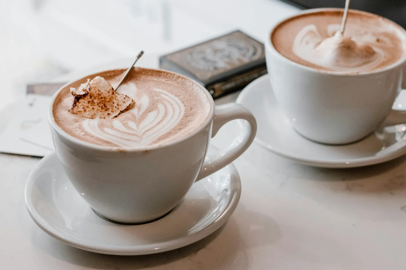 Duas xícaras brancas de café com leite sobre pires, em uma mesa clara de mármore